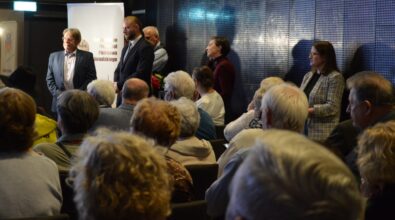 Na zdjęciu widać zgromadzoną publiczność siedzącą na krzesłach w sali spotkań, a także kilka osób stojących w przejściu między rzędami. Uczestnicy wydarzenia rozmawiają i przemieszczają się, co sugeruje zakończenie spotkania lub przerwę podczas wydarzenia odbywającego się w bibliotece.