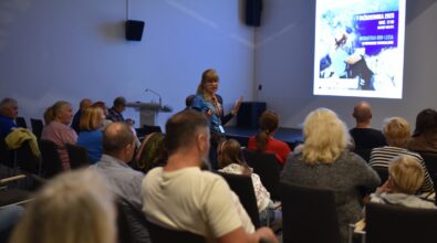 Wiesława Brudniak z Klubu Zdobywców Korony Gór Polski stoi między rzędami krzeseł w sali konferencyjnej i prowadzi wystąpienie, gestykulując podczas rozmowy z publicznością. Uczestnicy w różnym wieku siedzą zwróceni w jej stronę i uważnie słuchają prelekcji. W tle widoczny jest ekran z wyświetloną prezentacją dotyczącą spotkania w Mediatece 800-lecia, a całość odbywa się w nowoczesnej, dobrze oświetlonej przestrzeni sprzyjającej wydarzeniom edukacyjnym.