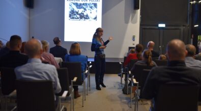 Uczestnicy siedzą na widowni sali konferencyjnej i słuchają prelekcji, a na ekranie wyświetlona jest plansza z logo Klubu Zdobywców Korony Gór Polski. Na pierwszym planie widoczna jest przedstawicielka klubu stojąca tyłem do aparatu, ubrana w koszulkę z nazwą organizacji, zwrócona w stronę publiczności i ekranu. Wnętrze sali jest nowoczesne, z rzędami krzeseł, oświetleniem sufitowym i skupioną na prezentacji publicznością.