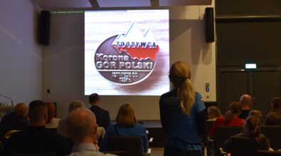 Uczestnicy siedzą na widowni sali konferencyjnej i słuchają prelekcji, a na ekranie wyświetlona jest plansza z logo Klubu Zdobywców Korony Gór Polski. Na pierwszym planie widoczna jest przedstawicielka klubu stojąca tyłem do aparatu, ubrana w koszulkę z nazwą organizacji, zwrócona w stronę publiczności i ekranu. Wnętrze sali jest nowoczesne, z rzędami krzeseł, oświetleniem sufitowym i skupioną na prezentacji publicznością.