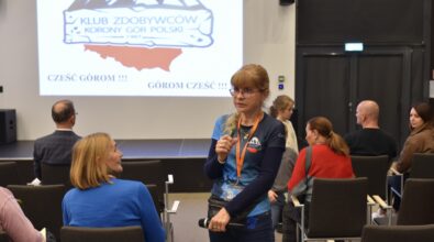 Wiesława Brudniak z Klubu Zdobywców Korony Gór Polski Wiesława Brudniak stoi w sali konferencyjnej i rozmawia z uczestnikami spotkania, trzymając w dłoni mikrofon. W tle widoczny jest ekran z wyświetlonym logo Klubu Zdobywców Korony Gór Polski oraz hasłem „Cześć górom! Górom cześć!”. Uczestnicy siedzą na krzesłach, część z nich odwraca się w stronę prelegentki, a w sali panuje atmosfera rozmowy i podsumowania spotkania.