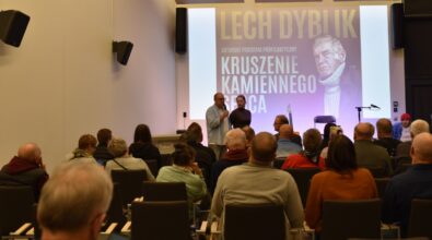 Publiczność zgromadzona w auli, w tle na ekranie widać plakat promujący spotkanie. Mężczyzna i kobieta stoją przed sceną, mężczyzna przemawia do mikrofonu.