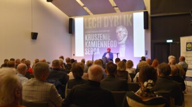 Lech Dyblik siedzi na scenie, obok niego gitara na stojaku i projekcja wizualna w tle, przed nim zgromadzona publiczność.