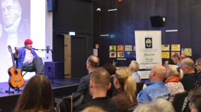 Lech Dyblik siedzi na scenie, obok niego gitara na stojaku i projekcja wizualna w tle, przed nim zgromadzona publiczność.