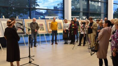 Na zdjęciu widoczna jest grupa uczestników zgromadzona w przestrzeni wystawowej podczas wydarzenia kulturalnego, stojąca przed prezentowanymi planszami z pracami. W centralnej części kadru znajduje się mikrofon na statywie, a cała scena dokumentuje moment oficjalnego spotkania towarzyszącego otwarciu wystawy.