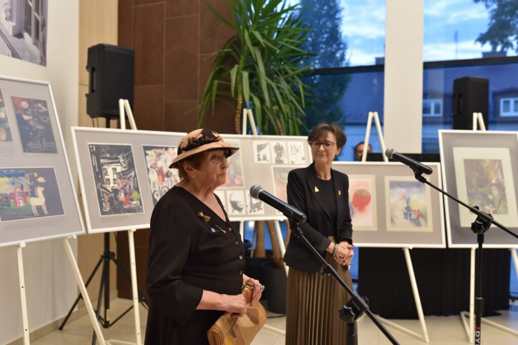 Dyrektor MBP i autorka wystawy stoją przy mikrofonach. W tle widać prace na sztalugach.