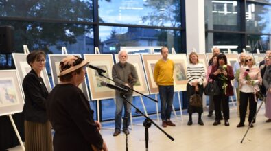 Na zdjęciu widoczna jest artystka stojąca przy mikrofonie i przemawiająca do zgromadzonych gości podczas otwarcia wystawy. W tle znajdują się uczestnicy wydarzenia oraz plansze z prezentowanymi pracami, a cała scena dokumentuje oficjalny moment spotkania kulturalnego.