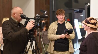 Na zdjęciu widoczny jest operator kamery rejestrujący przebieg wydarzenia oraz młody mężczyzna stojący obok z aparatem fotograficznym w dłoniach. Kadr dokumentuje medialną obsługę otwarcia wystawy i podkreśla profesjonalny charakter wydarzenia kulturalnego.