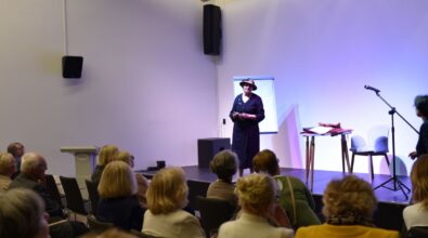 Na zdjęciu widoczna jest artystka stojąca na scenie i przemawiająca do zgromadzonej publiczności podczas spotkania autorskiego. Uczestnicy siedzą na sali i uważnie słuchają, a kadr oddaje spokojną, refleksyjną atmosferę wydarzenia kulturalnego.