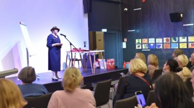 Na zdjęciu widoczna jest artystka stojąca przy mównicy i czytająca fragmenty tekstu podczas spotkania autorskiego. Przed sceną siedzi publiczność, a w tle widoczne są elementy scenografii oraz prezentowane prace, co podkreśla literacko-artystyczny charakter wydarzenia.