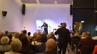 Na zdjęciu widoczna jest artystka stojąca na scenie i wykonująca gest podczas spotkania autorskiego, zwrócona w stronę zgromadzonej publiczności. Na sali siedzą uczestnicy wydarzenia, a w centralnej części kadru znajduje się operator kamery dokumentujący przebieg spotkania.