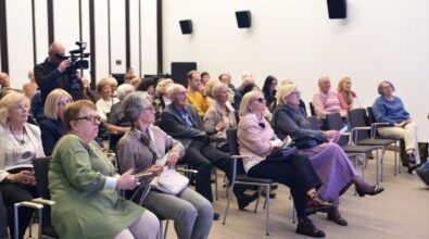 Na zdjęciu widoczna jest licznie zgromadzona publiczność siedząca na sali podczas otwarcia wystawy Agnieszki Illinicz-Kiełkiewicz „I tak to w życiu (u)bywa...”, w pierwszym rzędzie siedzi pani dyrektor Miejskiej Biblioteki Publicznej. Uczestnicy spotkania uważnie słuchają opowieści artystki o swojej twórczości.