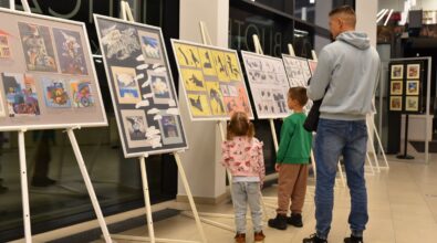 Na zdjęciu widoczne są dzieci wraz z opiekunem oglądające plansze wystawy ustawione na stojakach w przestrzeni biblioteki. Najmłodsi z zainteresowaniem przyglądają się ilustracjom, a kadr podkreśla rodzinny i międzypokoleniowy charakter wydarzenia kulturalnego.