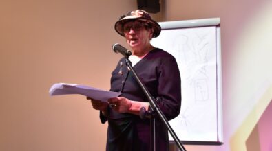 Na zdjęciu widoczny jest mężczyzna stojący przy mikrofonie i czytający tekst z kartki podczas spotkania. W tle znajduje się ekran z projekcją, a kadr oddaje oficjalny i literacki charakter wydarzenia kulturalnego.