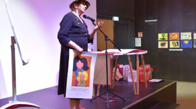 Na zdjęciu widoczna jest Agnieszka Illinicz-Kiełkiewicz stojąca na scenie przy mikrofonie podczas spotkania autorskiego. Obok znajduje się stolik z materiałami, a w tle widoczne są prace prezentowane w ramach wystawy, co podkreśla artystyczny charakter wydarzenia.