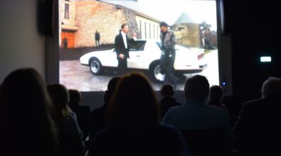 Grupa osób siedzi w ciemnym pomieszczeniu, oglądając film wyświetlany na dużym ekranie.