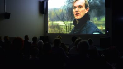 Grupa osób siedzi w ciemnym pomieszczeniu, oglądając film wyświetlany na dużym ekranie.