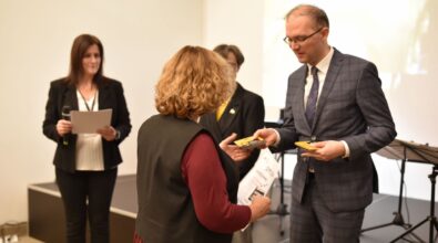 Na zdjęciu widoczny jest moment wręczania nagrody podczas uroczystego wydarzenia, w którym mężczyzna w garniturze przekazuje upominek kobiecie stojącej przodem do aparatu. W tle znajdują się inne osoby zaangażowane w organizację wydarzenia, a cała scena ma oficjalny charakter i podkreśla rangę gali finałowej.