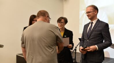 Na zdjęciu widać mężczyznę odbierającego dyplom lub nagrodę od przedstawicieli organizatorów wydarzenia stojących przed nim. Osoby wręczające dokumenty uśmiechają się, a całość przedstawia oficjalny moment podsumowania konkursu lub wydarzenia kulturalnego odbywającego się w bibliotece.