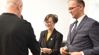 Na zdjęciu widać moment wręczania nagrody lub dyplomu uczestnikowi wydarzenia przez kobietę i mężczyznę stojących obok siebie. Organizatorzy uśmiechają się i przekazują upominek w jasnej sali, co wskazuje na oficjalne podsumowanie konkursu lub wydarzenia kulturalnego odbywającego się w bibliotece.