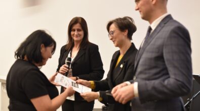 Na zdjęciu widać moment wręczania dyplomu lub nagrody uczestniczce wydarzenia przez przedstawicieli organizatorów stojących w jasnej sali. Kobieta odbiera dokument, a obok niej stoją osoby trzymające mikrofon i upominki, co wskazuje na oficjalne podsumowanie konkursu lub wydarzenia kulturalnego odbywającego się w bibliotece.