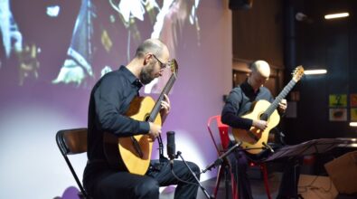 Dwóch mężczyzn siedzi na scenie i gra na gitarach klasycznych. Przed nimi stoją pulpity muzyczne i mikrofony. W tle widać projekcję filmową.