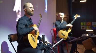 Na zdjęciu widać dwóch muzyków siedzących na krzesłach i grających na gitarach podczas kameralnego koncertu w przestrzeni bibliotecznej. Artyści występują na tle ekranu projekcyjnego, a całość sugeruje wydarzenie muzyczne lub koncert towarzyszący spotkaniu kulturalnemu w Mediatece.