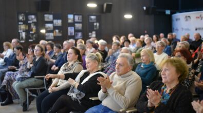 Na zdjęciu widoczna jest licznie zgromadzona publiczność siedząca na sali podczas wydarzenia kulturalnego i nagradzająca występujących brawami. Uczestnicy w różnym wieku wypełniają przestrzeń, a w tle widoczne są ściany z prezentowanymi fotografiami, co podkreśla uroczysty i wspólnotowy charakter spotkania.
