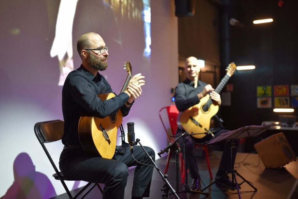 Dwóch mężczyzn siedzi na scenie i gra na gitarach klasycznych. Przed nimi stoją pulpity muzyczne i mikrofony. W tle widać projekcję filmową.
