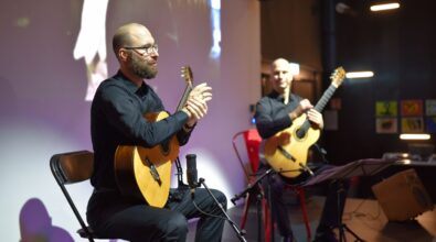 Dwóch mężczyzn siedzi na scenie i gra na gitarach klasycznych. Przed nimi stoją pulpity muzyczne i mikrofony. W tle widać projekcję filmową.