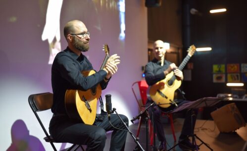 Dwóch mężczyzn siedzi na scenie i gra na gitarach klasycznych. Przed nimi stoją pulpity muzyczne i mikrofony. W tle widać projekcję filmową.