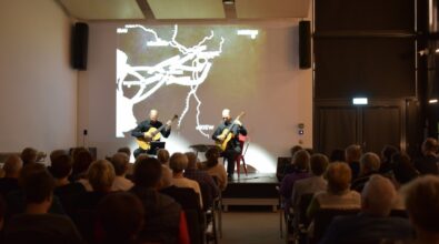 Na zdjęciu widoczna jest scena koncertowa z dwoma gitarzystami występującymi przed licznie zgromadzoną publicznością siedzącą na widowni. W tle znajduje się duży ekran z dynamiczną wizualizacją, a całość oddaje atmosferę koncertu muzyki filmowej będącego finałem projektu kulturalnego.