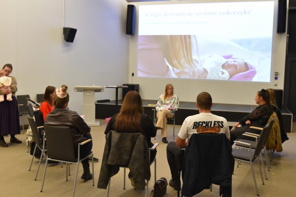 Zdjęcie przedstawia spotkanie edukacyjne w sali Mediateki, w którym uczestniczy grupa dorosłych osób siedzących w kręgu na krzesłach. W centrum znajduje się położna prowadząca rozmowę z uczestnikami, a spotkanie ma formę otwartej dyskusji. W tle widoczny jest ekran z prezentacją multimedialną, na której wyświetlone jest zdjęcie noworodka oraz slajd z pytaniem zachęcającym do refleksji i rozmowy. Atmosfera spotkania jest kameralna i sprzyjająca wymianie doświadczeń, a część uczestników trzyma notatniki lub siedzi z dziećmi.