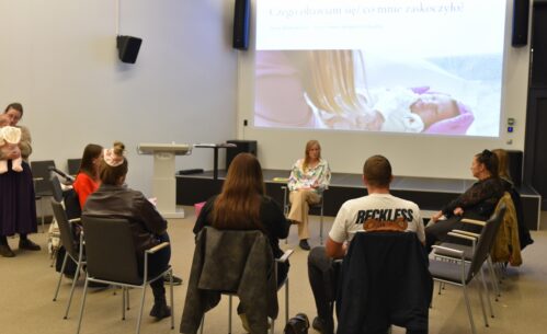 Zdjęcie przedstawia spotkanie edukacyjne w sali Mediateki, w którym uczestniczy grupa dorosłych osób siedzących w kręgu na krzesłach. W centrum znajduje się położna prowadząca rozmowę z uczestnikami, a spotkanie ma formę otwartej dyskusji. W tle widoczny jest ekran z prezentacją multimedialną, na której wyświetlone jest zdjęcie noworodka oraz slajd z pytaniem zachęcającym do refleksji i rozmowy. Atmosfera spotkania jest kameralna i sprzyjająca wymianie doświadczeń, a część uczestników trzyma notatniki lub siedzi z dziećmi.