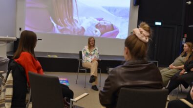 Zdjęcie przedstawia kameralne spotkanie edukacyjne w sali Mediateki, podczas którego uczestnicy siedzą w kręgu na krzesłach i słuchają prowadzącej rozmowę kobiety siedzącej na środku. W tle widoczny jest duży ekran z prezentacją multimedialną, na którym wyświetlone jest zdjęcie noworodka. Uczestnicy zwróceni są w stronę prowadzącej, a atmosfera spotkania ma charakter spokojnej, otwartej rozmowy sprzyjającej zadawaniu pytań i wymianie doświadczeń.