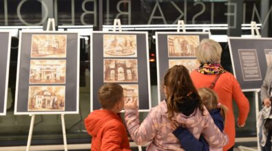 Dzieci wraz z dorosłymi oglądają plansze z grafikami podczas otwarcia wernisażu w Mediatece. Uczestnicy stoją przed ekspozycją i przyglądają się przedstawionym widokom architektury. Prace zaprezentowane są na sztalugach w przeszklonej przestrzeni biblioteki. Zdjęcie ukazuje międzypokoleniowy charakter wydarzenia i wspólne odkrywanie wystawy.