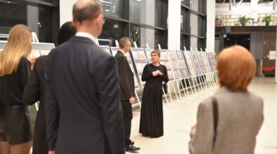 Grupa osób uczestniczy w otwarciu wernisażu w przestrzeni wystawowej Mediateki. Kuratorka lub prowadząca wydarzenie zwraca się do zgromadzonych, stojąc na tle plansz ekspozycyjnych ustawionych w długim rzędzie. Uczestnicy słuchają i oglądają prezentowane prace w nowoczesnym, przeszklonym wnętrzu biblioteki. Zdjęcie dokumentuje oficjalny moment inauguracji wystawy.