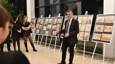 Mężczyzna w garniturze zwraca się do zgromadzonych podczas otwarcia wernisażu, stojąc na tle plansz z prezentowanymi pracami. Za nim widoczna jest ekspozycja grafik ustawionych na sztalugach w długim rzędzie. Obok stoją uczestniczki wydarzenia, słuchające wystąpienia. Zdjęcie dokumentuje oficjalny moment inauguracji wystawy w przestrzeni Mediateki.