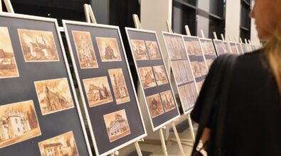 Na zdjęciu widoczne są plansze wystawowe ustawione na sztalugach, prezentujące prace graficzne w ciepłej, sepiowej kolorystyce. Grafiki przedstawiają architekturę i pejzaże miejskie. Przed planszami stoi uczestniczka wernisażu, oglądająca ekspozycję. Zdjęcie ukazuje fragment wystawy prezentowanej podczas otwarcia w Mediatece.