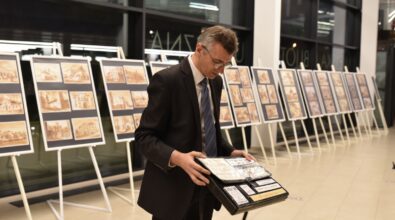 Mężczyzna w garniturze stoi w przestrzeni wystawowej i przegląda teczkę z materiałami. W tle ustawione są plansze ekspozycyjne na sztalugach z ilustracjami i grafikami. Wnętrze Mediateki jest jasne i nowoczesne, z dużymi przeszkleniami. Zdjęcie dokumentuje przygotowania lub zwiedzanie wystawy.