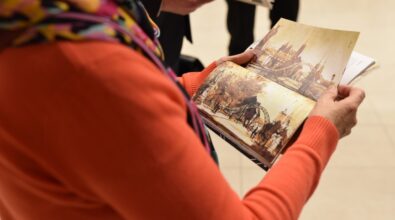 Osoba trzyma w dłoniach otwartą książkę i ogląda ilustracje przedstawiające miejskie pejzaże. W tle widoczne są dłonie innych uczestników z publikacjami, co sugeruje wspólne przeglądanie materiałów. Zdjęcie ukazuje moment skupienia i zainteresowania treścią książek. Całość oddaje kameralną, spokojną atmosferę spotkania.