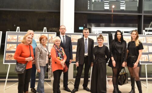 Grupa uczestników stoi do wspólnego zdjęcia podczas otwarcia wernisażu, na tle plansz z prezentowanymi pracami. Wśród zgromadzonych widoczni są goście, organizatorzy oraz osoby zaangażowane w przygotowanie wystawy. Ekspozycja umieszczona na sztalugach wypełnia przestrzeń Mediateki. Zdjęcie dokumentuje oficjalny moment inauguracji wydarzenia i spotkanie uczestników.