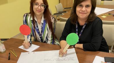 Zdjęcie przedstawia dwie kobiety siedzące przy stole w sali szkoleniowej lub konferencyjnej. Obie mają na szyjach identyfikatory i trzymają w dłoniach kolorowe tabliczki na patykach, jedną czerwoną i jedną zieloną, wykorzystywane podczas ćwiczeń lub warsztatów. Na stole leżą kartki papieru, długopisy oraz notatki, co wskazuje na pracę warsztatową lub szkoleniową. Kobiety uśmiechają się do obiektywu, a atmosfera spotkania ma charakter edukacyjny i angażujący.