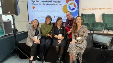 Zdjęcie przedstawia grupę pięciu osób siedzących i stojących na scenie konferencyjnej przed dużym ekranem z planszą zatytułowaną „Konferencja Społeczeństwo i Nauka. Zaufanie – to skomplikowane?”, organizowaną przez Centrum Popularyzacji Nauki i Innowacji Kortosfera. Uczestnicy mają na szyjach identyfikatory konferencyjne, co wskazuje na udział w wydarzeniu naukowym lub edukacyjnym. Osoby ubrane są w stroje o charakterze półformalnym, a całość utrzymana jest w profesjonalnej, konferencyjnej atmosferze. W tle widoczne są elementy sceny, nagłośnienie oraz logotypy organizatorów i partnerów wydarzenia.