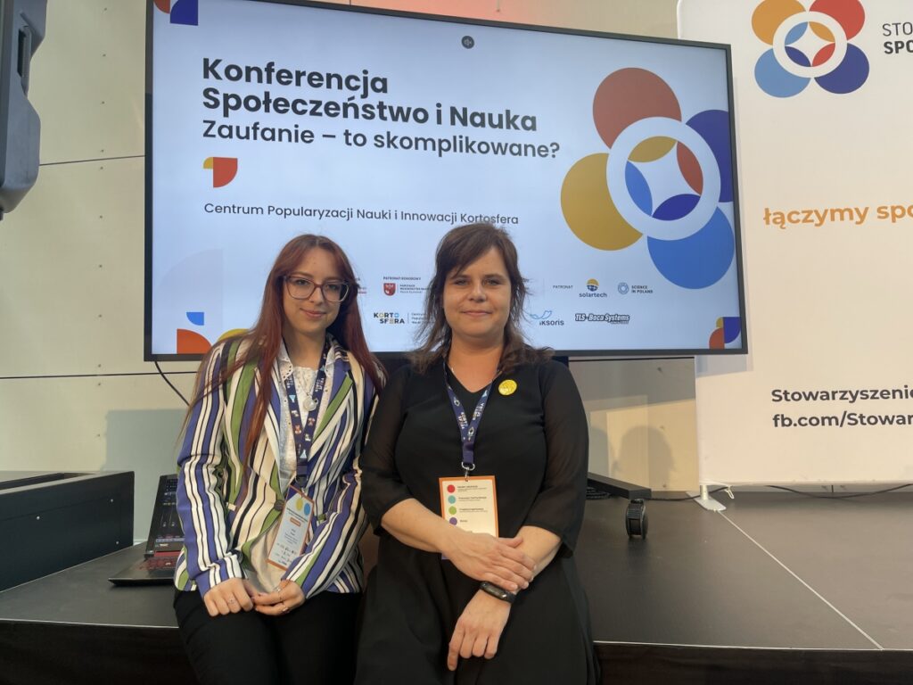 Zdjęcie przedstawia dwie kobiety stojące obok siebie przed dużym ekranem z planszą konferencyjną zatytułowaną „Konferencja Społeczeństwo i Nauka. Zaufanie – to skomplikowane?”, organizowaną przez Centrum Popularyzacji Nauki i Innowacji Kortosfera. Obie kobiety mają na szyjach identyfikatory konferencyjne, co wskazuje na udział w wydarzeniu naukowym lub edukacyjnym. Jedna z nich ubrana jest w jasną koszulę i pasiastą marynarkę, druga w czarną sukienkę z przypinką. Tło stanowi scena konferencyjna z logotypami partnerów i instytucji, a całość ma formalny, profesjonalny charakter.