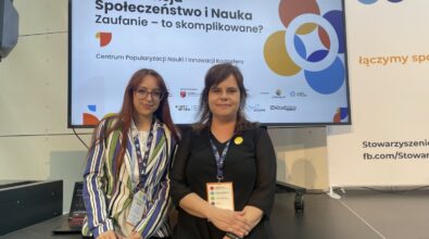 Zdjęcie przedstawia dwie kobiety stojące obok siebie przed dużym ekranem z planszą konferencyjną zatytułowaną „Konferencja Społeczeństwo i Nauka. Zaufanie – to skomplikowane?”, organizowaną przez Centrum Popularyzacji Nauki i Innowacji Kortosfera. Obie kobiety mają na szyjach identyfikatory konferencyjne, co wskazuje na udział w wydarzeniu naukowym lub edukacyjnym. Jedna z nich ubrana jest w jasną koszulę i pasiastą marynarkę, druga w czarną sukienkę z przypinką. Tło stanowi scena konferencyjna z logotypami partnerów i instytucji, a całość ma formalny, profesjonalny charakter.