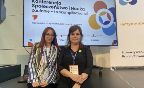 Zdjęcie przedstawia dwie kobiety stojące obok siebie przed dużym ekranem z planszą konferencyjną zatytułowaną „Konferencja Społeczeństwo i Nauka. Zaufanie – to skomplikowane?”, organizowaną przez Centrum Popularyzacji Nauki i Innowacji Kortosfera. Obie kobiety mają na szyjach identyfikatory konferencyjne, co wskazuje na udział w wydarzeniu naukowym lub edukacyjnym. Jedna z nich ubrana jest w jasną koszulę i pasiastą marynarkę, druga w czarną sukienkę z przypinką. Tło stanowi scena konferencyjna z logotypami partnerów i instytucji, a całość ma formalny, profesjonalny charakter.