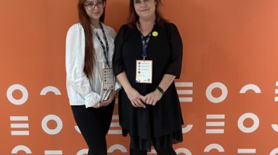 Zdjęcie przedstawia dwie kobiety stojące obok siebie na tle pomarańczowej ścianki z logotypem Uniwersytetu Warmińsko-Mazurskiego w Olsztynie. Obie mają na szyjach identyfikatory konferencyjne, co wskazuje na udział w wydarzeniu naukowym lub edukacyjnym. Jedna z kobiet ubrana jest w jasną bluzkę i ciemne spodnie, druga w czarną sukienkę i czarne buty na grubej podeszwie. Kobiety stoją swobodnie, zwrócone przodem do obiektywu, a całość utrzymana jest w formalno-konferencyjnym charakterze.