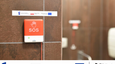 Na zdjęciu widoczny jest czerwony przycisk alarmowy SOS zamontowany na ścianie w toalecie dostępnej w Mediatece. Urządzenie wyposażone jest w oznaczenia wizualne oraz elementy w alfabecie Braille’a, co umożliwia jego obsługę także osobom z niepełnosprawnością wzroku. System przywoławczy pozwala na szybkie wezwanie pomocy w sytuacji wymagającej wsparcia, zwiększając bezpieczeństwo oraz dostępność przestrzeni dla wszystkich użytkowników biblioteki. Na fotografii widoczne są również oznaczenia informujące o realizacji projektu w ramach Funduszy Europejskich dla Rozwoju Społecznego.