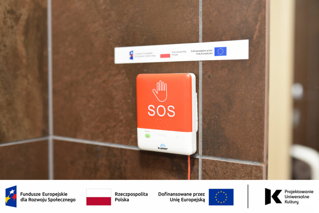 Na zdjęciu widoczny jest czerwony przycisk alarmowy SOS zamontowany na ścianie w toalecie dostępnej w Mediatece. Urządzenie wyposażone jest w czytelne oznaczenia graficzne oraz elementy w alfabecie Braille’a, dzięki czemu może być używane także przez osoby z niepełnosprawnością wzroku. System przywoławczy umożliwia szybkie wezwanie pomocy w sytuacji wymagającej wsparcia, zwiększając bezpieczeństwo i dostępność przestrzeni dla wszystkich użytkowników biblioteki. Na fotografii widoczne są również oznaczenia informujące o realizacji projektu w ramach Funduszy Europejskich dla Rozwoju Społecznego oraz programu „Projektowanie Uniwersalne Kultury”.